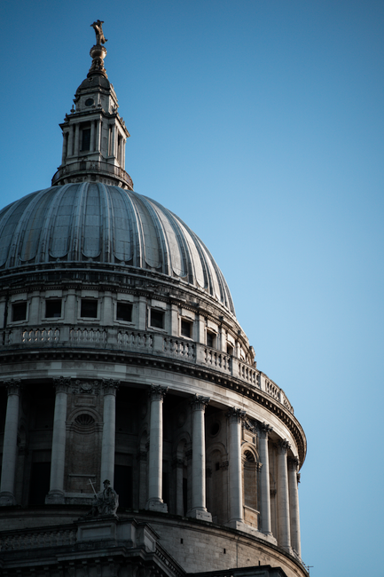 Aerial view of St. Paul's Cathedral in London, showing its iconic large stone dome with decorative columns and lantern at the top. The surrounding cityscape includes modern and historic buildings with