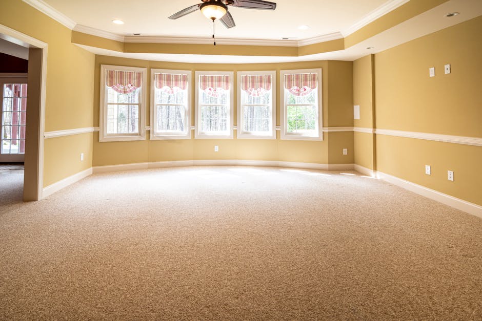 A spacious living room with a beige carpeted floor that appears clean and well-maintained, reflecting light from five large windows with white frames and pink-striped valances. The walls are painted i