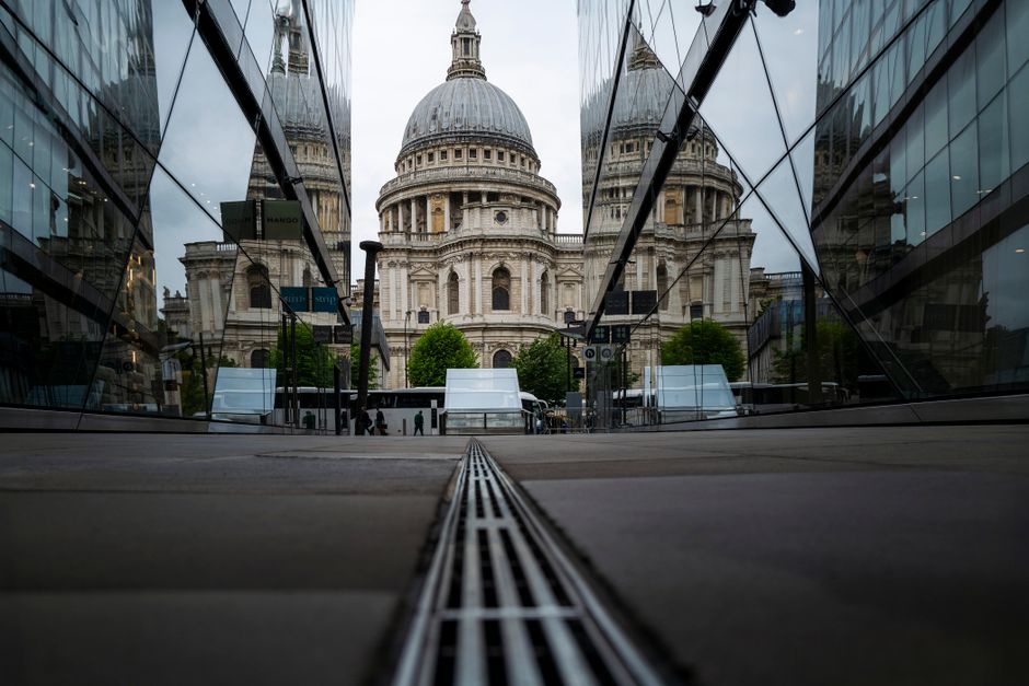 A low-angle view of St Paul's Cathedral in London framed by modern glass buildings on either side, with reflective surfaces showing the surrounding cityscape. The scene shows a paved pedestrian area w