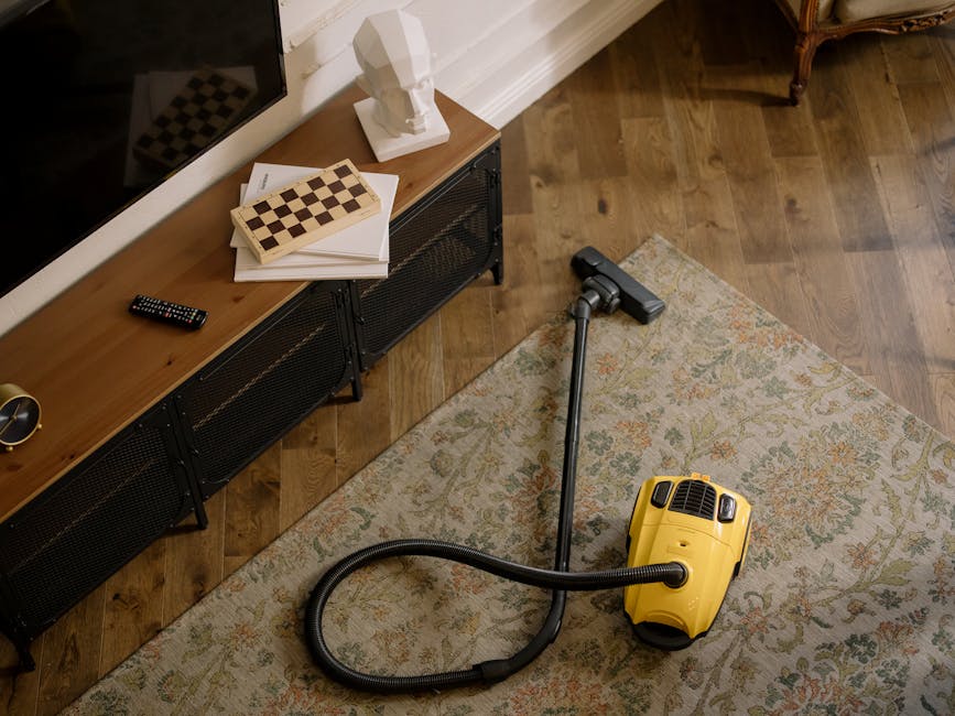 A yellow handheld vacuum cleaner with a flexible hose and cleaning head lying on a patterned area rug in a living room. The room features a wooden floor, a black media console with metal mesh doors, a