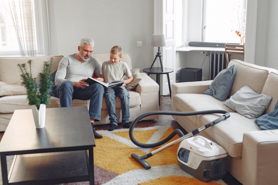 A yellow handheld vacuum cleaner with a flexible hose and cleaning head lying on a patterned area rug in a living room. The room features a wooden floor, a black media console with metal mesh doors, a